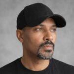 Portrait of Duane Whitfield wearing a black baseball cap and black shirt against a light gray background.